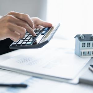 Person using calculator with paperwork and small house model, suggesting home financial planning